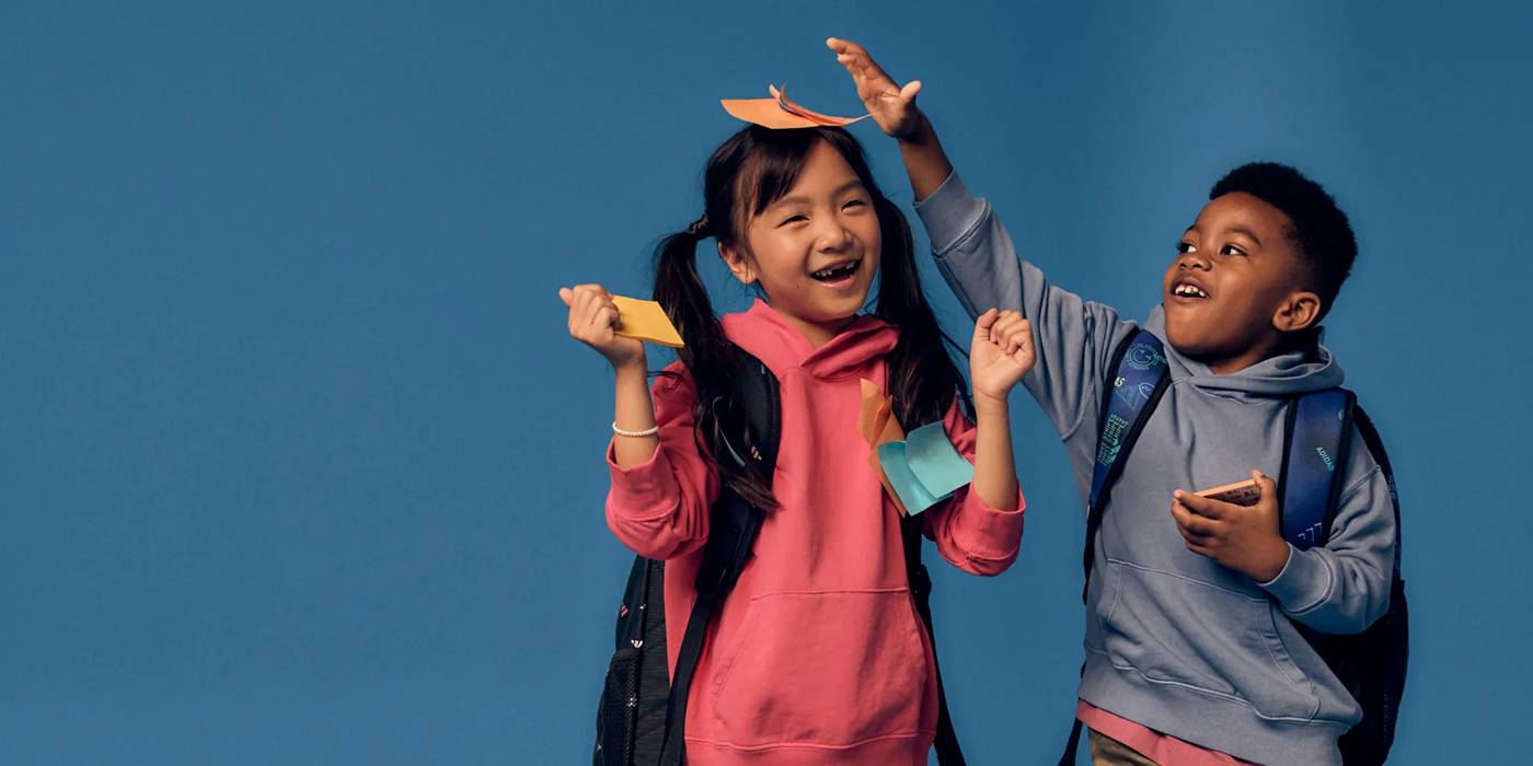 Two school-aged children wearing backpacks, smiling and placing sticky notes on one another.
